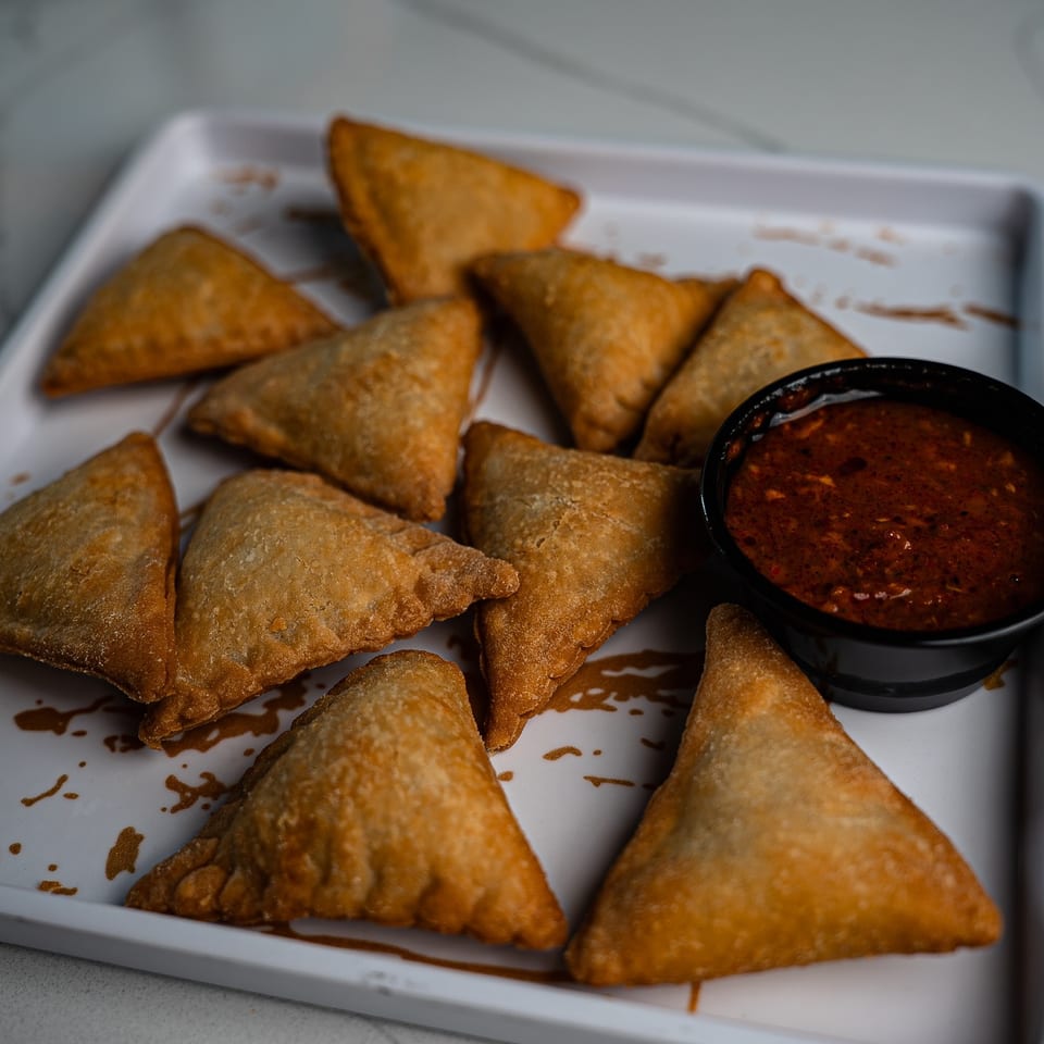 Golden fried samosas arranged on a white platter beside a bowl of spicy red dipping sauce.