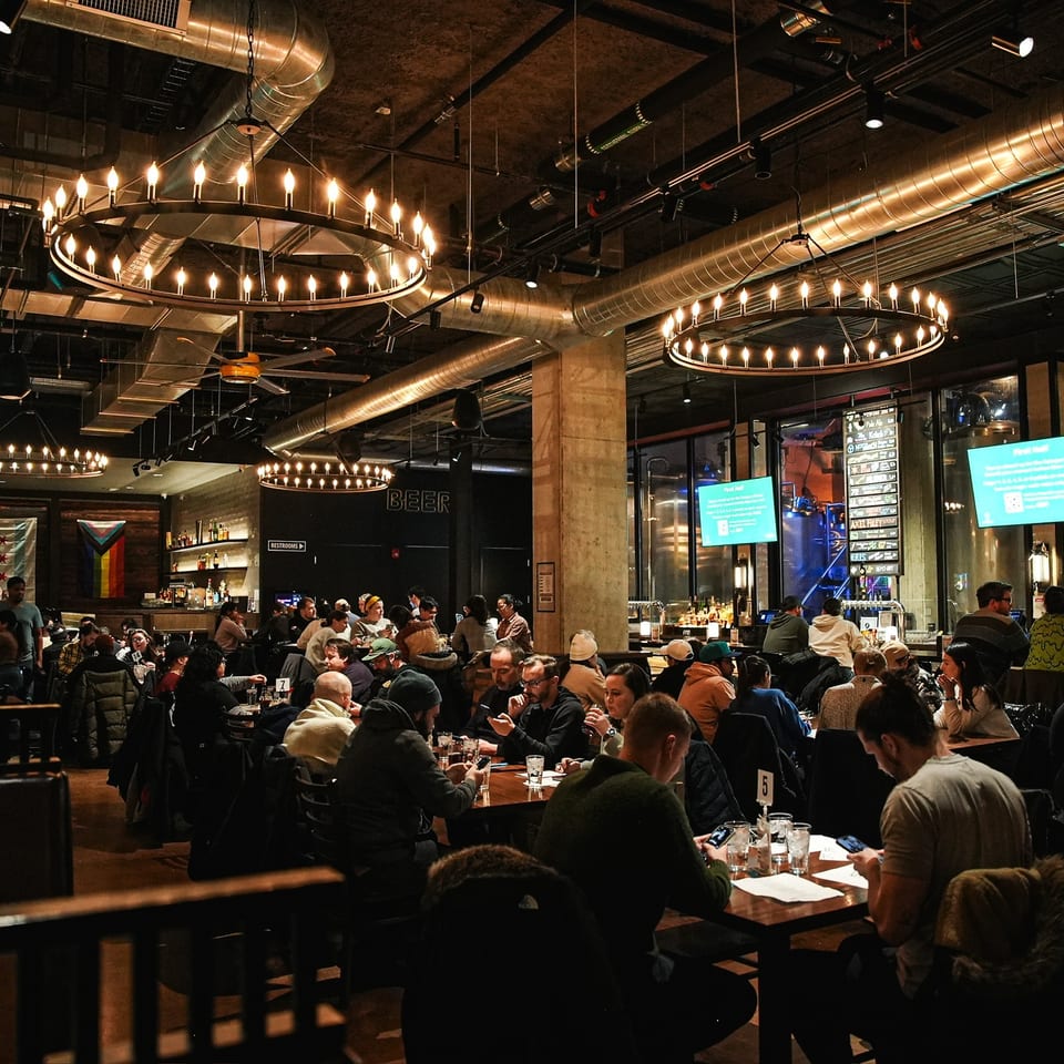 Busy restaurant dining room with guests at tables, a large bar, exposed ductwork, and circular candle-style chandeliers.