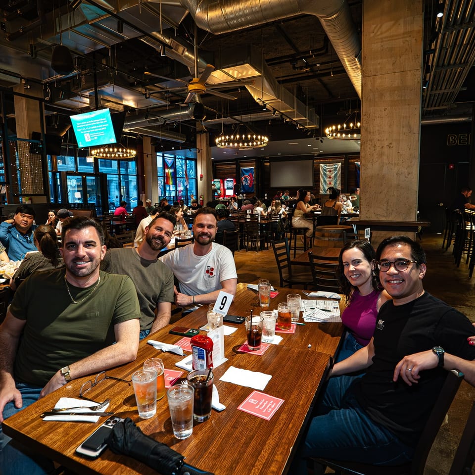 Friends smile around a restaurant table with drinks beneath glowing chandeliers and exposed industrial ductwork.