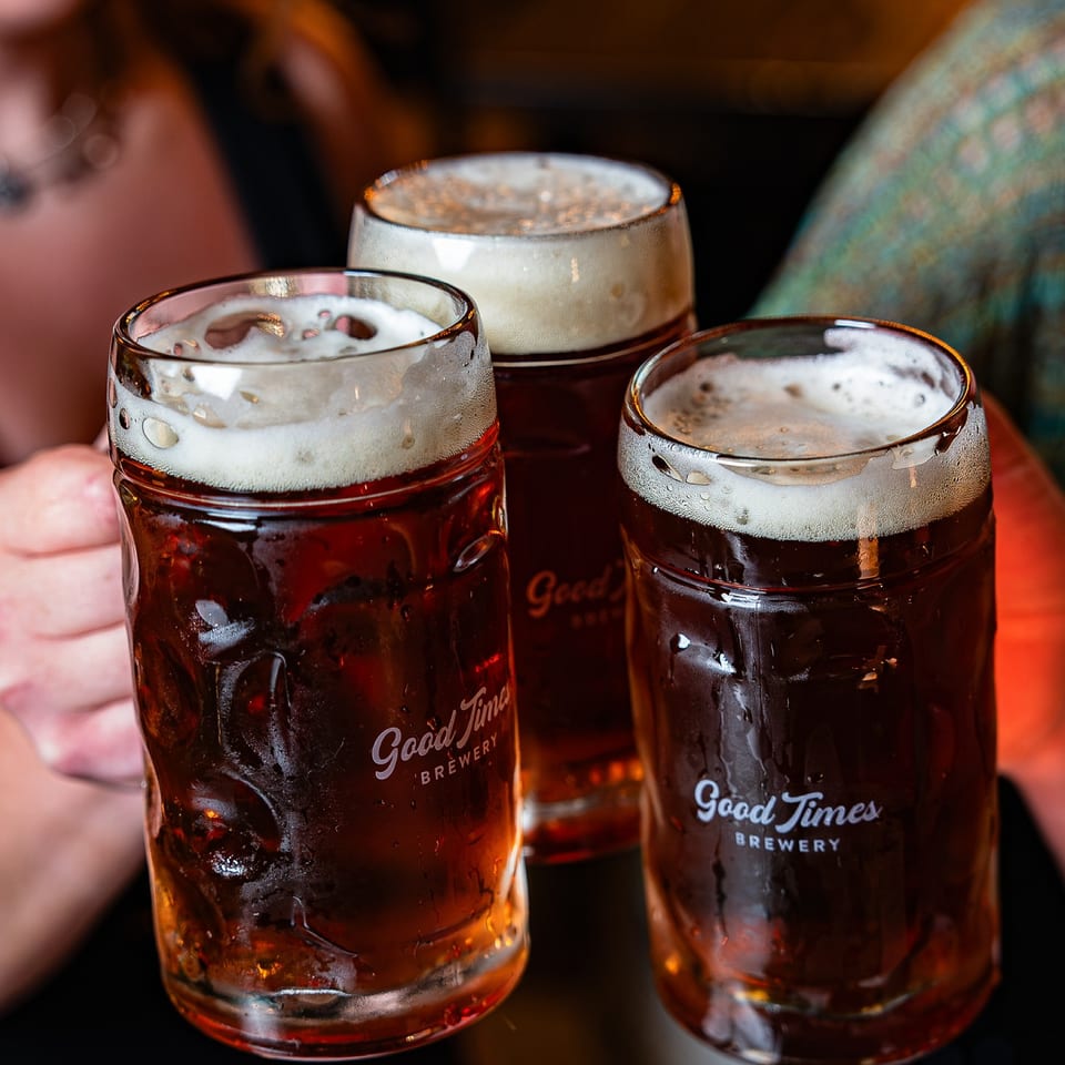 Three frothy dark beers in branded Good Times Brewery glasses on a warmly lit restaurant table, with people blurred behind them.