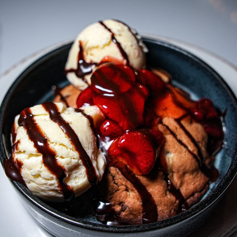 Black bowl filled with vanilla and chocolate ice cream, strawberries, cookies, and chocolate sauce.