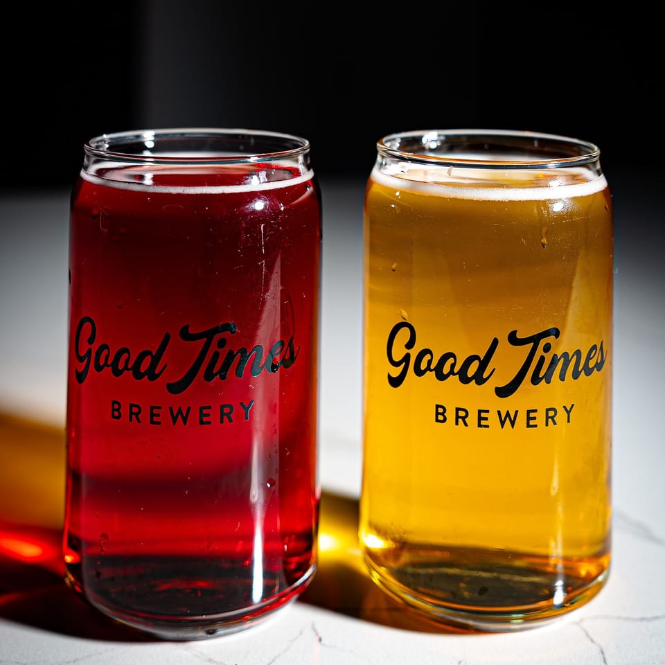Two Good Times Brewery glasses hold red and golden beers on a countertop against a dark background.