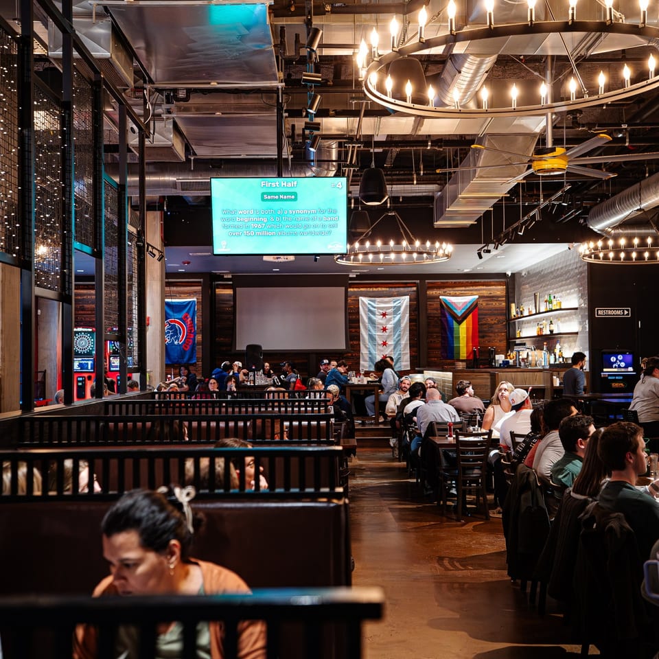 Busy sports restaurant with diners, arcade games, large trivia screen, exposed ductwork, and illuminated circular chandeliers.