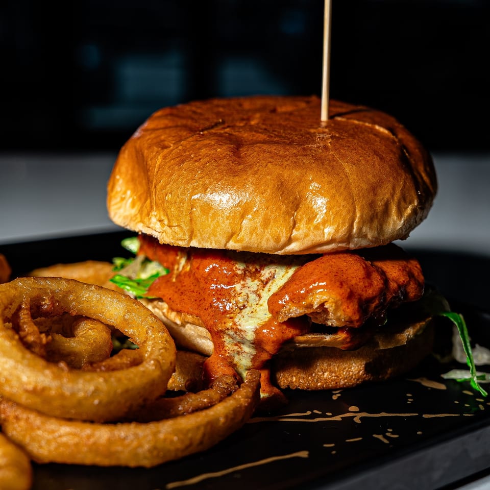 Gourmet burger with a glossy brioche bun, saucy fried chicken, greens, and onion rings on a black serving plate.