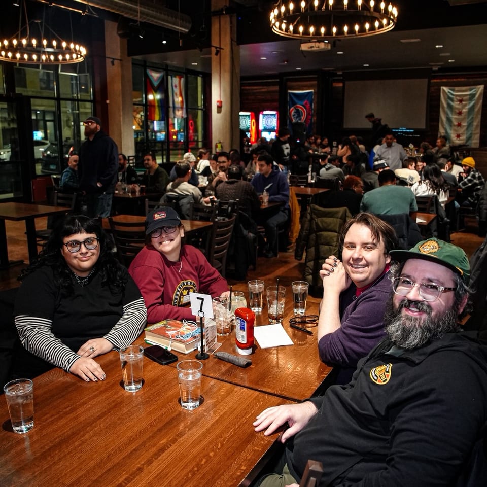 Friends gather around a restaurant table with drinks beneath glowing chandeliers in a busy, warmly lit dining room.