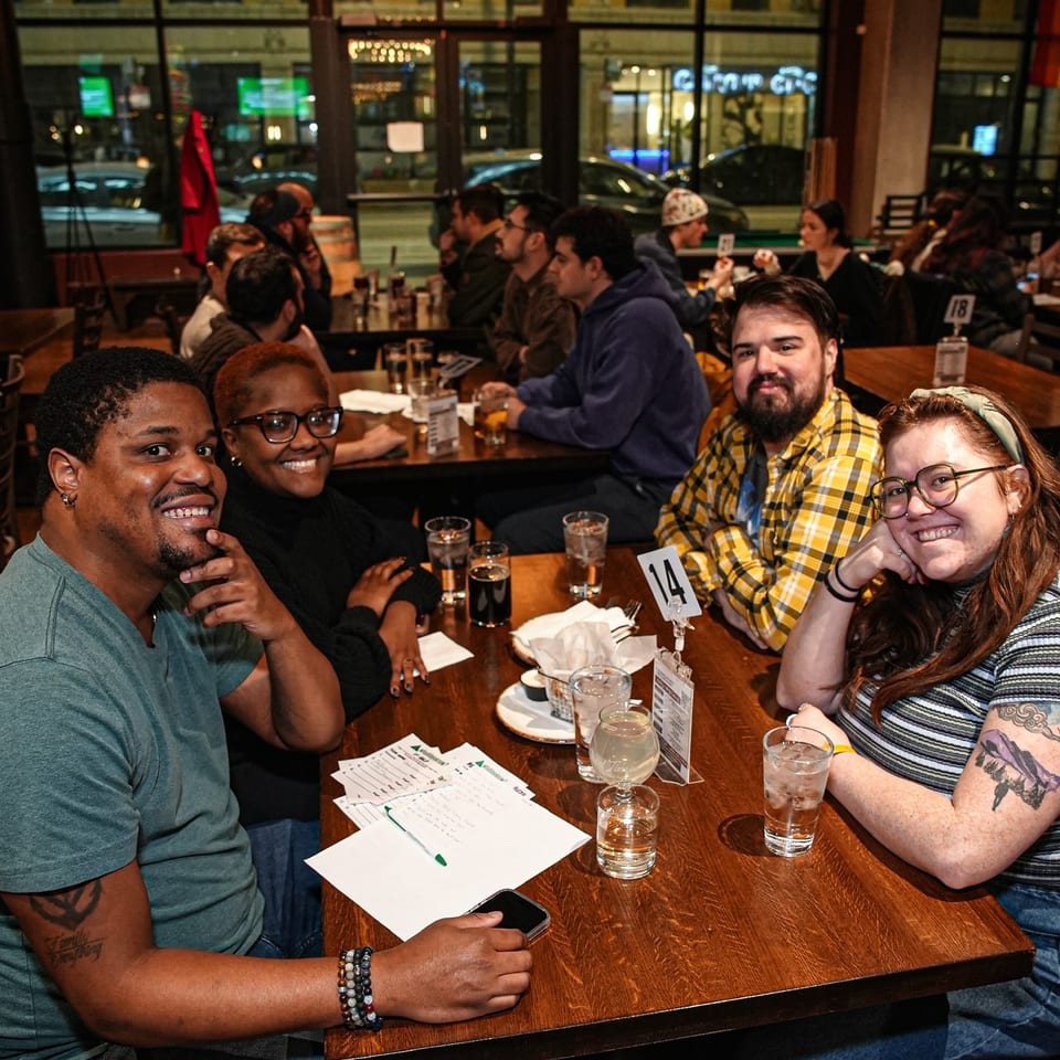 Friends smile around a restaurant table with drinks and papers beneath a glowing circular chandelier.