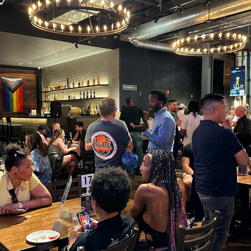 Busy sports bar dining room with diverse patrons, tables, exposed ducts, glowing chandeliers, and LGBTQ-themed flags.