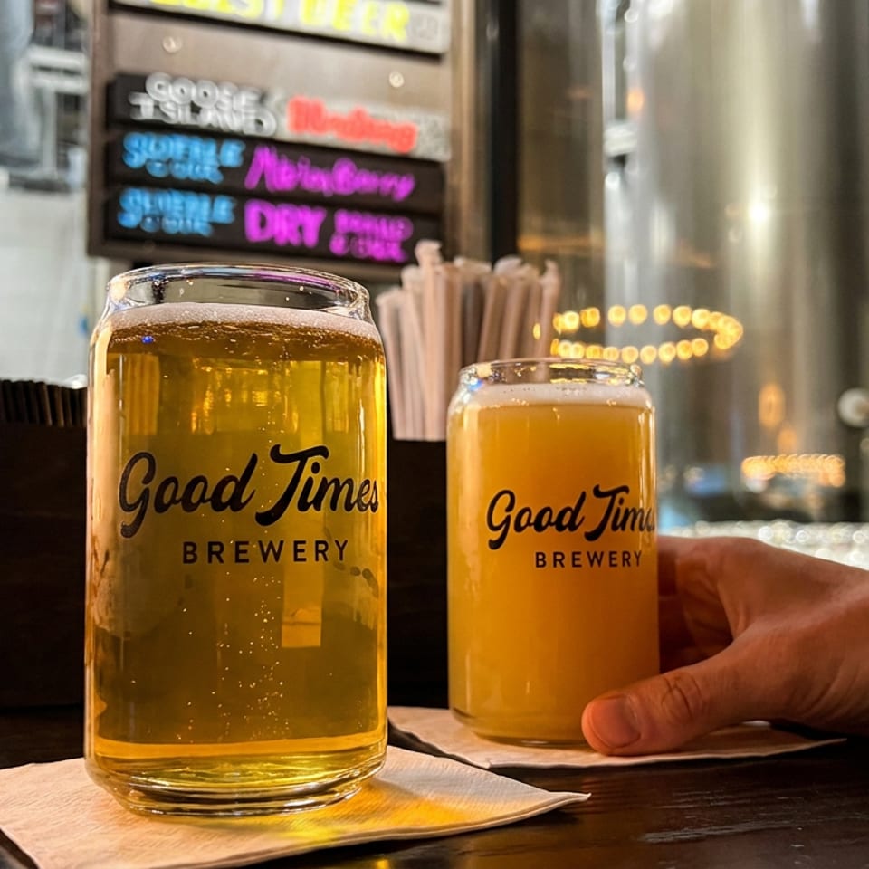 Two Good Times Brewery beers sit on napkins at a warmly lit bar, with a hand reaching toward the hazy orange beer.
