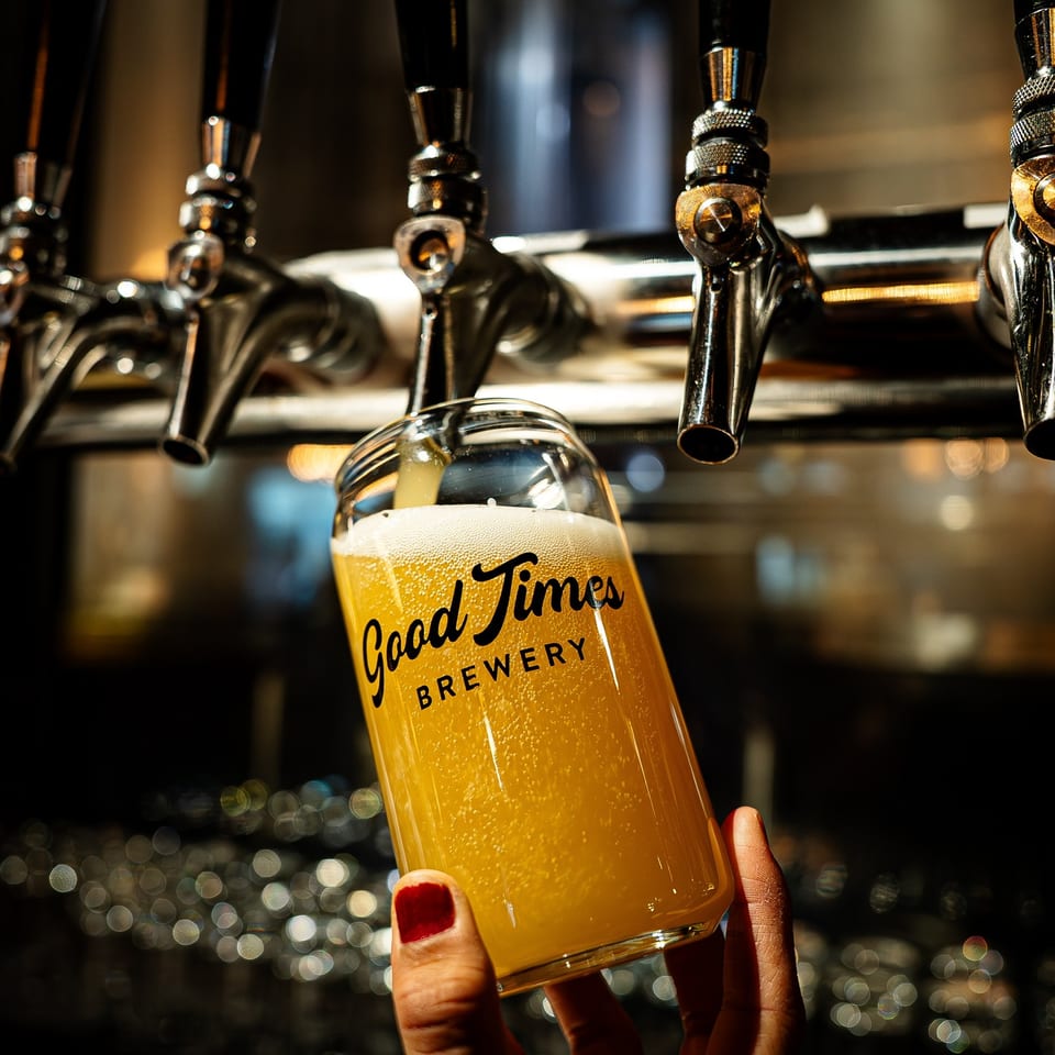 Hand holding a glass of golden beer labeled Good Time Brewery beneath illuminated bar taps.