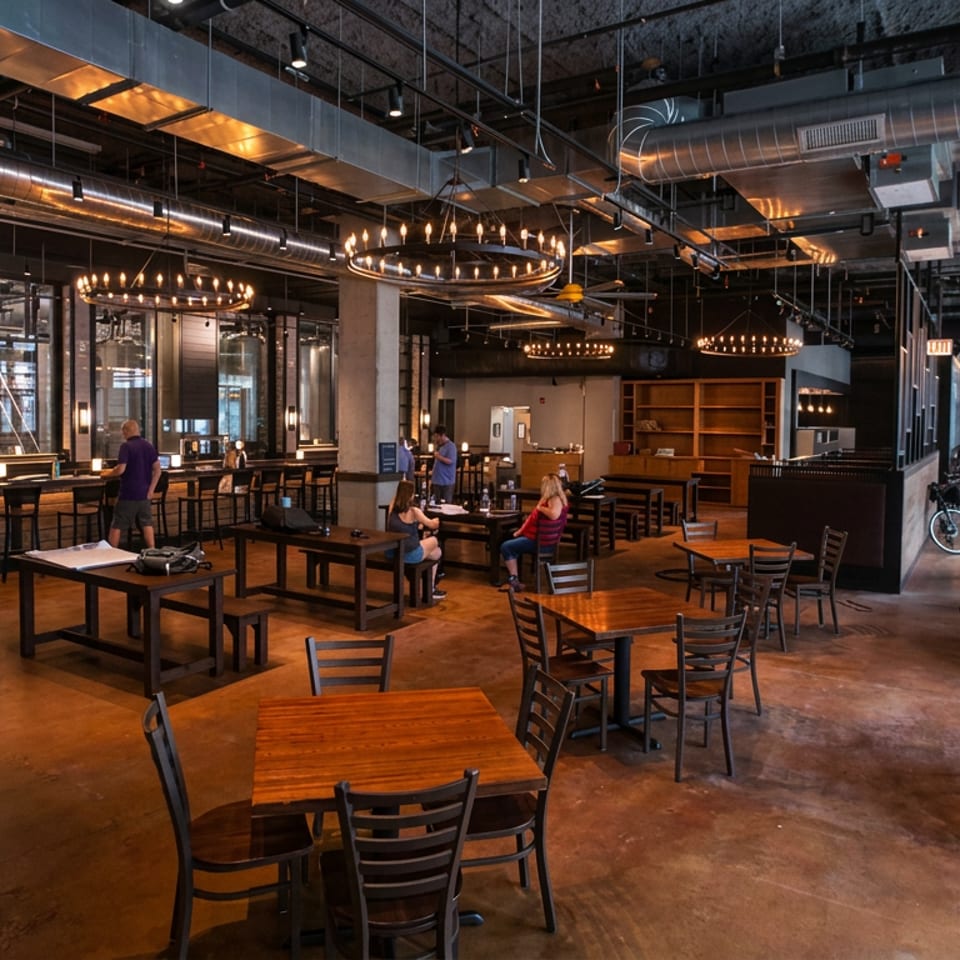 Spacious industrial-style restaurant interior with exposed ductwork, warm pendant lighting, wooden tables, bar seating, and several patrons.