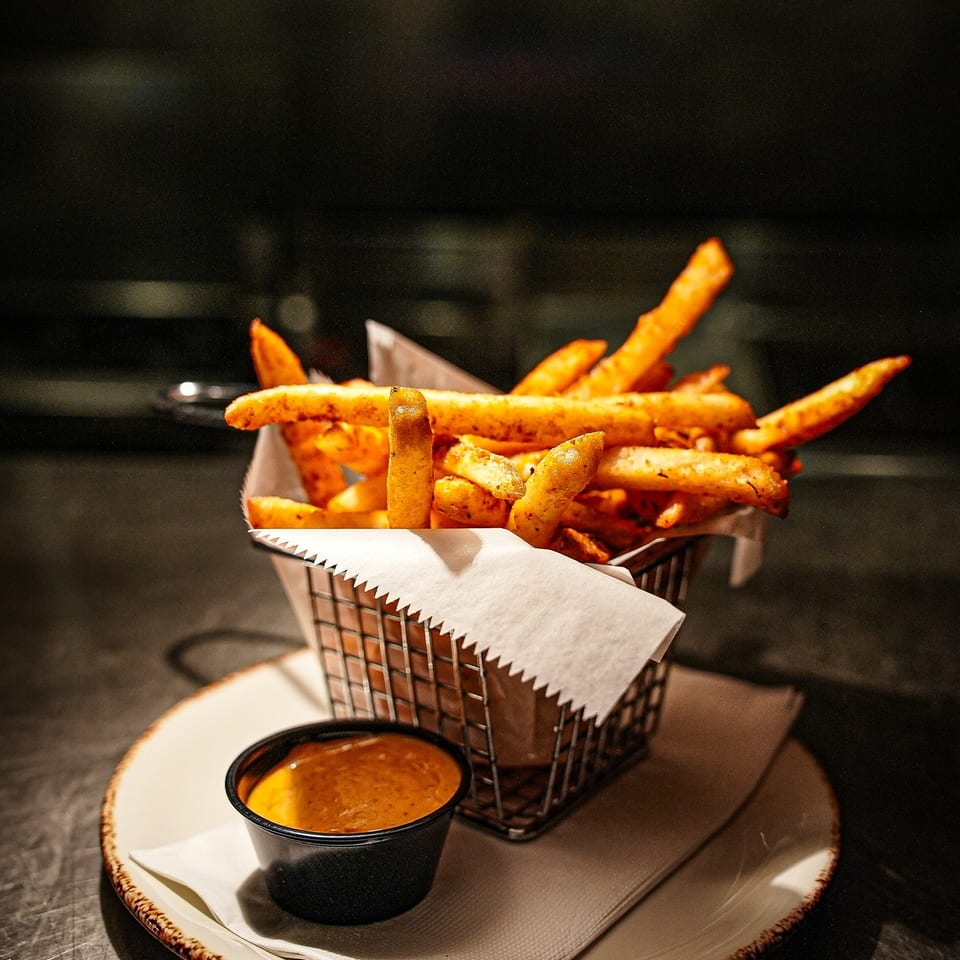Crispy golden fries fill a wire basket on a plate, served with a cup of dipping sauce.