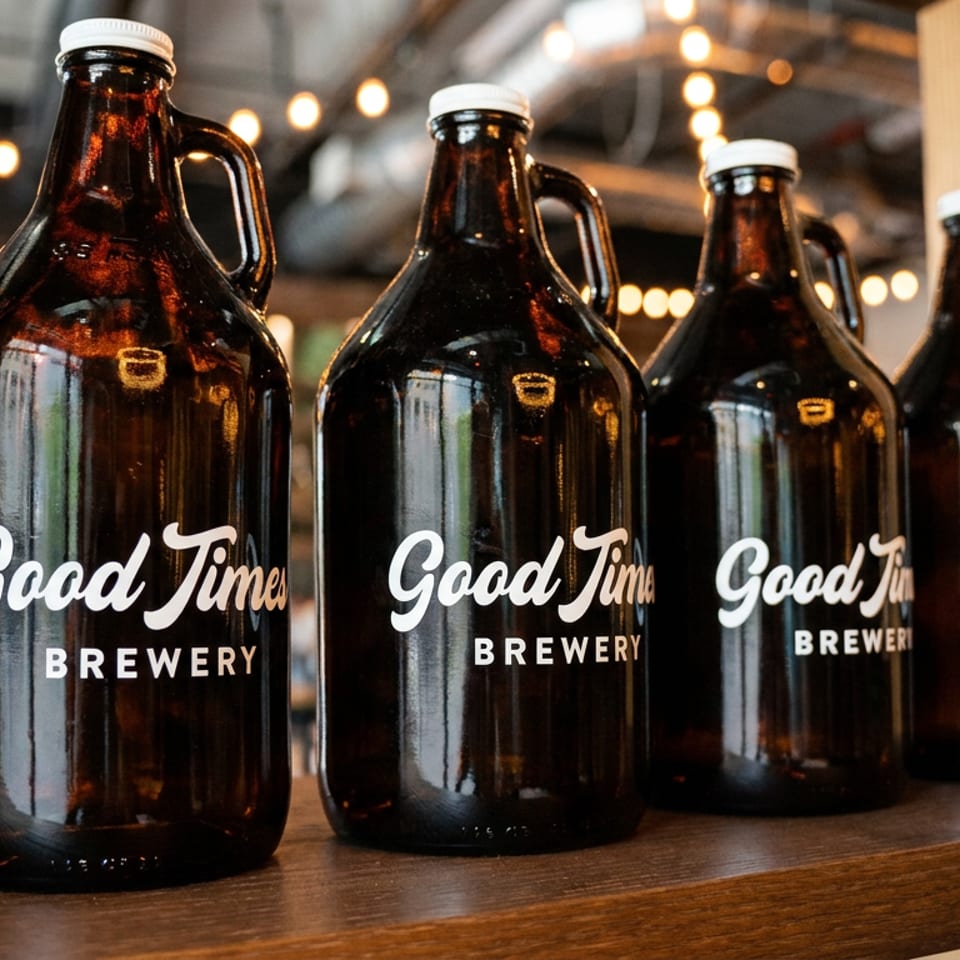 Row of brown Good Times Brewery growlers displayed on a wooden shelf beneath warm pendant lights.