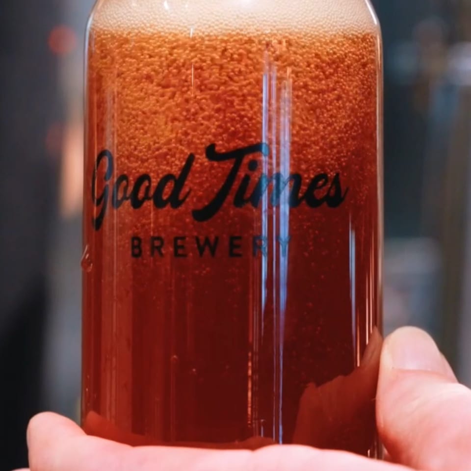 A hand holds a glass of amber Good Times Brewing beer beneath a tap, topped with thick white foam.