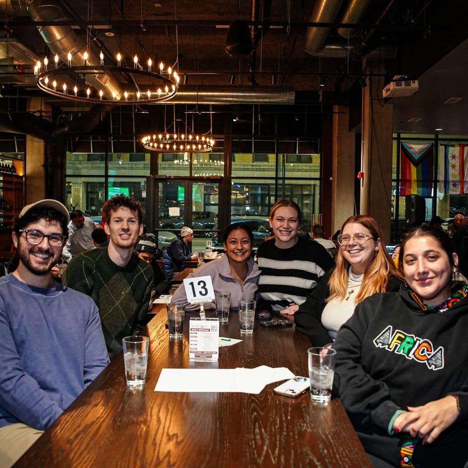Six friends smile around a restaurant table beneath glowing circular chandeliers, with trivia materials and drinks on the table.