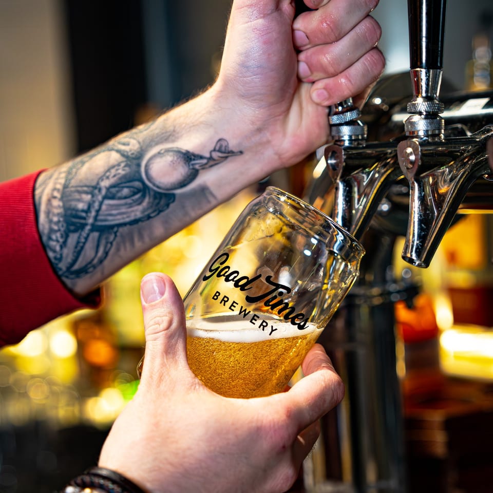 Bartender pours golden beer from a tap into a branded glass at a warmly lit brewery bar.