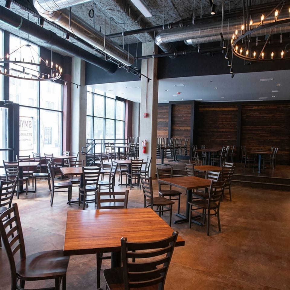 Spacious industrial-style restaurant dining room with wooden tables, black chairs, exposed ductwork, and circular chandeliers.